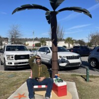 Palm tree sculpture rises from a cluster of sculpted Adirondack chairs arranged in a circle, painted in soft pastel shades of blue and pink. Positioned on a plaza with patterned stone tiles, the artwork blends casual beach vibes with playful design in the heart of downtown Columbia, SC.