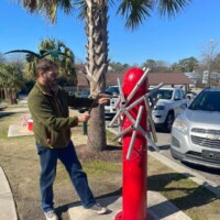 Man standing beside a tall red musical sculpture with angled metal rods in Columbia, SC; framed by palm trees, parked cars, and the city’s urban backdrop