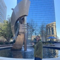 Abstract metal sculpture with curved and angular shapes rises from the center of a circular fountain, surrounded by a brick border. The sculpture features interlocking loops and arcs, reflecting light and adding kinetic energy to the urban plaza. In the background, modern glass architecture contrasts with classical building facades and leafy trees, blending Columbia’s evolving skyline with public art.
