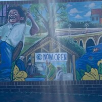 Vibrant mural in Columbia, SC painted on a brick wall, showing two children on swings to the left—one in a yellow shirt, the other in white—suspended in mid-air with joyful expressions. Centered is a small house labeled “Home 09,” beneath a rainbow and fluffy clouds. To the right, a stone bridge arches over a river flanked by yellow flowers, leading into a cityscape of colorful buildings. A person in a green jacket stands beside the mural, adding scale and realism.