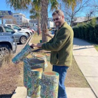 Person playing a set of colorful outdoor drum sculptures in Columbia, SC, each painted with abstract patterns in orange, blue, and red. The instruments are placed along a sunny sidewalk next to a parking lot, with palm trees and parked cars in the background, blending public art with playful sound.