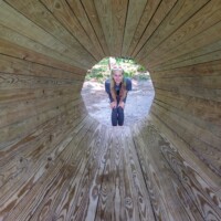 Person framed inside circular wooden tunnel at Agnes Andreae Nature Preserve Michigan — radial plank design, forest light, smiling pose