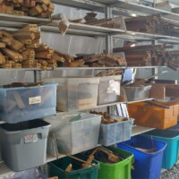 Organized shelving with stacked wooden blocks, planks, and labeled plastic bins filled with assorted wood pieces inside a reuse workshop.