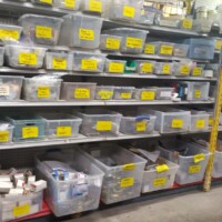 Shelving unit filled with clear plastic bins labeled with yellow tags, each containing organized electronic components, tools, and small parts in a reuse workshop.