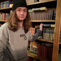 Person in black beanie and gray sweatshirt standing in front of a large bookshelf with ornate spines, captured during the Ghosts & Gravestones tour in a vintage study setting.