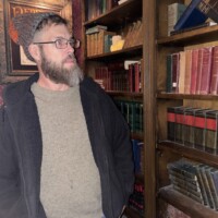 Person in knit hat and glasses standing in front of a wooden bookshelf filled with colorful books, captured during the Ghosts & Gravestones tour in a warmly lit vintage study.