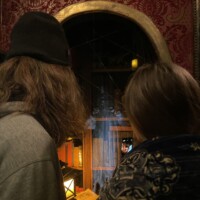 Two people facing a dimly lit alcove with golden arches and red wallpaper, one wrapped in a patterned blanket and the other holding up a phone, with a holographic ghost floating between them.