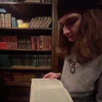 Person in black beanie and gray sweatshirt reading an open book in front of a vintage bookshelf, captured during the Ghosts & Gravestones tour in a dimly lit study.