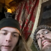 Two people posing for a selfie in front of red and gold patterned curtains, one wearing a black hat with an orange band, the other in a knit cap and glasses, under warm indoor lighting.