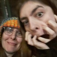 Two people posing for a selfie in a dark setting, one wearing a colorful knit hat and glasses, the other making a dramatic face with wide eyes and hands raised.