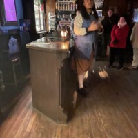Tour guide in colonial attire standing behind a candlelit counter, speaking to a small group inside a dimly lit room with wooden floors and vintage shelves.