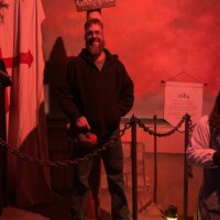 Man standing under a wooden “Garrote” sign holding a severed head prop, with a red cross banner and historical signage in the background, lit by lantern glow at the Ghost & Gravestones Tour St Augustine.