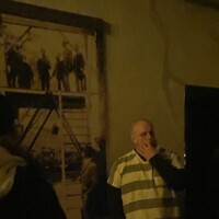 Tour guide in striped prison costume standing in front of a historical mural inside a dimly lit room, mid-conversation with a guest.