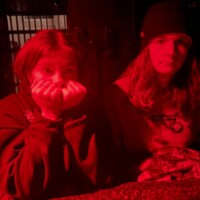 Two people sitting at a table under red lighting, one resting their face on their hands and the other wearing a beanie, with barred windows in the background.