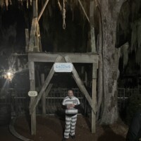 Tour guide in striped prison costume standing under a wooden gallows at night, with a noose overhead and trees in the background.