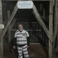 Tour guide in striped prison-style costume standing in front of a wooden gallows structure, part of the Ghosts and Gravestones tour St Augustine.
