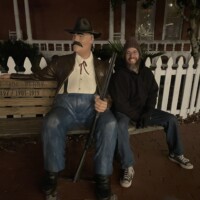 Person sitting next to a life-sized cowboy statue holding a rifle, in front of a white picket fence and a “City Hall” sign, under dim nighttime lighting at the Ghosts & Gravestones Tour St Augustine.