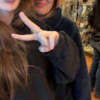 Two people posing for a selfie inside a souvenir shop, one making a peace sign and sticking out their tongue, the other smiling in a colorful knit hat, with arched windows and shelves in the background.