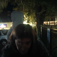 Person in a hoodie at night, with a statue looming behind and a lit-up building in the background—ghoul vibes fully intact.