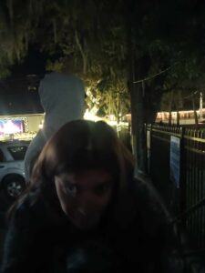 Person in a hoodie at night, with a statue looming behind and a lit-up building in the background—ghoul vibes fully intact.