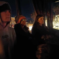 Three people sitting close together in a dimly lit bus, dressed in hoodies and knit hats, with holiday lights glowing outside the window.