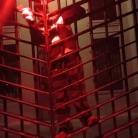 Tour guide in striped prison costume clinking metal bars on a jail cell under red lighting, with architectural details in the background.
