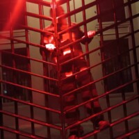 Tour guide in striped prison costume behind a metal jail cell under red lighting, with architectural details in the background.