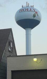 Holly Michigan water tower #3 with red lettering and holly leaf graphic, framed by rooftops and a glowing porch light under a clear evening sky.