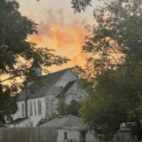 Downtown Holly Michigan near Battle Alley at sunrise, with church steeple, railroad tracks, and glowing orange sky creating a peaceful small-town atmosphere.
