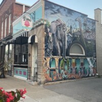 Colorful mural in downtown Holly Michigan featuring surreal elephant-headed figure, pyramids, and ‘TODAY’ text beside Zoup! storefront with striped awning