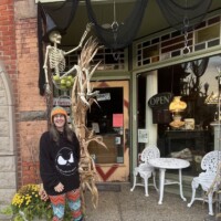 Downtown Holly Michigan storefront decorated for Halloween with skeleton among cornstalks, vintage architecture, and person in skull sweatshirt posing on sidewalk.