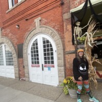 Downtown Holly Michigan building with arched white double doors, Halloween decorations including skeleton and cornstalks, and person in skull sweatshirt standing beside yellow flowers.