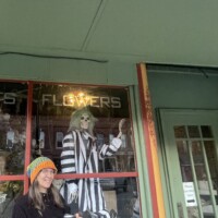 Downtown Holly Michigan storefront with green trim and Halloween window display featuring Beetlejuice figure and pumpkin, person in beanie holding coffee on sidewalk
