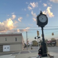 Downtown Holly Michigan street clock at sunset, with person seated on bench and warm light illuminating clouds and buildings in background.