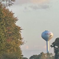 Holly Michigan water tower #2 with red lettering and holly leaf graphic, surrounded by fall foliage and trees under a partly cloudy sky