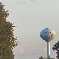 Holly Michigan water tower #2 with red lettering and holly leaf graphic, sun flare glowing through the berry under a partly cloudy sky.