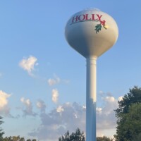 Holly Michigan water tower #2 with red lettering and holly leaf graphic, set against a partly cloudy sky and surrounded by trees.