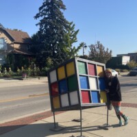 Outdoor sculpture of oversized Rubik’s Cube on metal frame in Holly Michigan with person leaning against it, street and houses in background