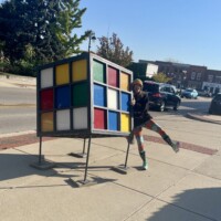 Oversized Rubik’s Cube sculpture on metal frame in Holly Michigan with person posing on one leg beside it, street, cars, and buildings in background.