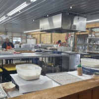 A commercial kitchen with stainless steel equipment and large prep tables. Two workers wearing aprons are preparing food. Containers of icing, trays, and baking supplies are spread across the tables. The space has bright overhead lighting and unfinished wood walls.