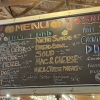 A large chalkboard menu hangs inside a market building. It lists food items such as soups, sandwiches, nachos, soft pretzels, mac and cheese, cider, and donuts. The board is handwritten in various colors and displayed above the counter, with wooden ceiling beams visible overhead.