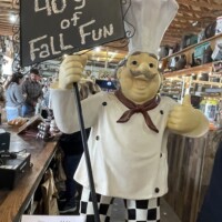 A mini life-size statue of a chef wearing a white coat, tall chef’s hat, and black-and-white checkered pants stands inside a market building. The chef is holding a chalkboard sign that reads “40 years of Fall Fun.” Baked goods in clear packaging are displayed on the counter in front of the statue, with shelves and overhead wooden rafters visible in the background.