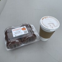 A takeout cup of hot cocoa sits next to a clear plastic container of chocolate-covered donuts on an outdoor picnic table. The container has a Johnson’s Pumpkin Farm label. The background is out of focus, showing open outdoor space.