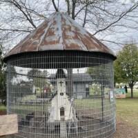 A round wire chicken coop with a rusted metal conical roof sits on a grassy area. Inside the coop is a small white wooden structure designed to look like a church. Trees and other farm buildings are visible in the background under an overcast sky.
