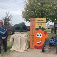 A person stands next to a fall-themed photo setup at Johnson’s Pumpkin Farm. The display includes a large Baby Yoda figure made from greenery, a height-measuring board with a smiling pumpkin graphic labeled “Johnson’s Pumpkin Farm 2023,” and a small blue tractor. A tree and fall decorations are in the background.