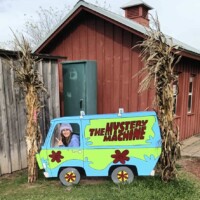A person poses behind a painted wooden cutout of the Mystery Machine van from Scooby-Doo, with their face visible through the driver’s window. The cutout is displayed outdoors between two cornstalk decorations, in front of a red wooden building with a small cupola on top.