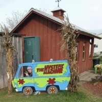 A person poses behind a painted wooden cutout of the Mystery Machine van from Scooby-Doo, with their face centered in the driver’s window and their feet hidden behind the board. The cutout is set outdoors between two cornstalk decorations, in front of a red wooden building with a small cupola on the roof.