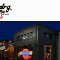 Exterior of The Laundry Restaurant in Fenton Michigan at night, with neon signage, large windows, and ‘25 Years’ anniversary banner reading ‘A Common Thread in the Village.