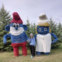 A woman stands between two large hay sculptures designed to resemble Papa Smurf and Smurfette. The figures are made from stacked hay bales painted blue and dressed with red pants and a white dress. Evergreen trees are behind them.