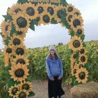 A person stands beneath a decorative archway covered in artificial sunflowers that reads “Johnson’s Sunflower Maze.” They are wearing a light blue hoodie, black skirt, and knit hat. Behind the arch is a field of sunflowers under an overcast sky, with straw bales on the ground in front.